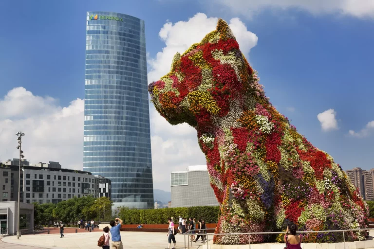 Bilbao, Spanien - August 30, 2013: Touristen fotografieren den großen Blumenhund "Puppy", der eingerahmt ist, als würde er ein Gebäude küssen; den Iberdrola-Wolkenkratzer