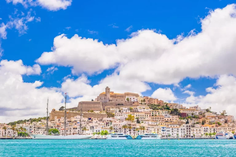 En ekstremt detaljeret udsigt over Eivissas gamle bymidte og marina, lys himmel, maleriske skyer, et fortøjet sejlskib, den ikoniske skyline i Dalt Vila domineret af katedralkirken Santa Maria de les Neus. Fremkaldt fra RAW.