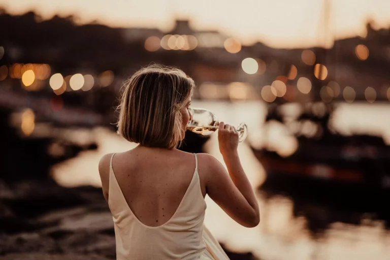 a lady in evening gown with a glass of white wine enjoying sunset sitting at a river Douro bank.